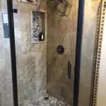 Modern walk-in shower with black frame enclosure, travertine tile walls, pebble stone floor, and built-in shelving with bathroom products