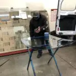 Worker in dark clothing and blue mask sitting at table next to white delivery van and stacked cardboard boxes